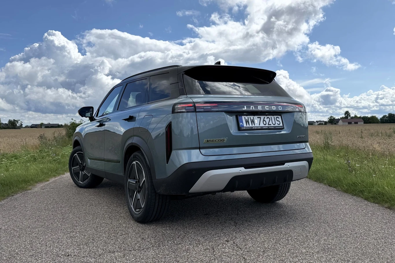 Jasnoniebieski SUV marki Jaecoo zaparkowany na asfaltowej drodze, otoczony polami i pod rozległym, częściowo pochmurnym niebem.