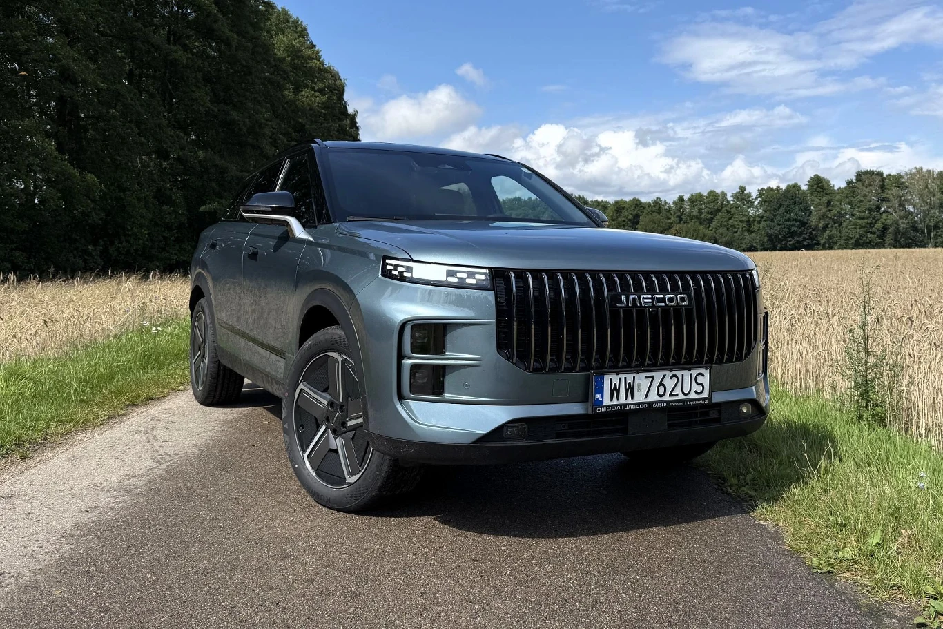 Srebrny SUV marki Jaecoo z dużym czarnym grillem stoi na wiejskiej drodze, obok pola uprawnego i zielonych drzew, pod jasnym, lekko zachmurzonym niebem.