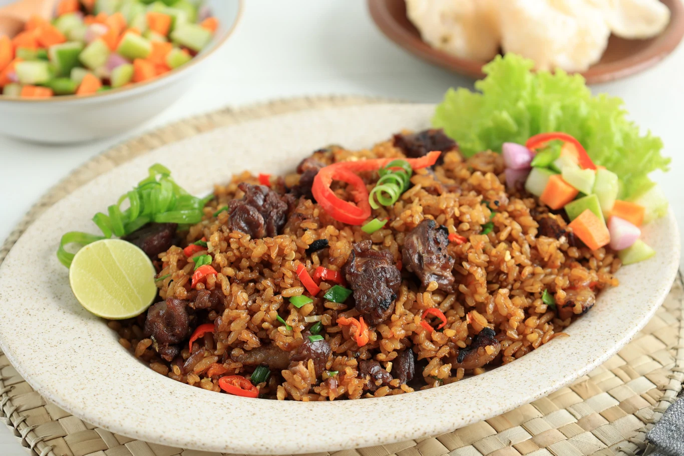Smak nasi goreng na długo pozostanie w pamięci Porcja indonezyjskiego nasi goreng z kawałkami mięsa, czerwoną papryką, szczypiorkiem, limonką i warzywami na sałacie.
