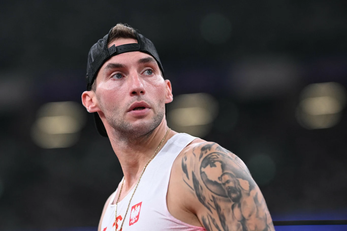 Marcin Krukowski Mężczyzna w sportowej koszulce z polskim godłem i czapce z daszkiem założonej tył na przód, z widocznym tatuażem na ramieniu, patrzy w bok na tle rozmytego stadionu.