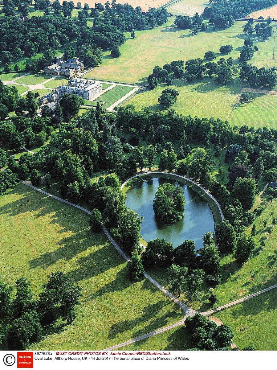 Miejsce pochówku księżnej Diany na jeziorze w Althorp House Duży pałac otoczony rozległymi ogrodami, na pierwszym planie owalny staw z wyspą porośniętą drzewami, wszystko otoczone zielenią i gęsto rozmieszczonymi drzewami parkowymi.