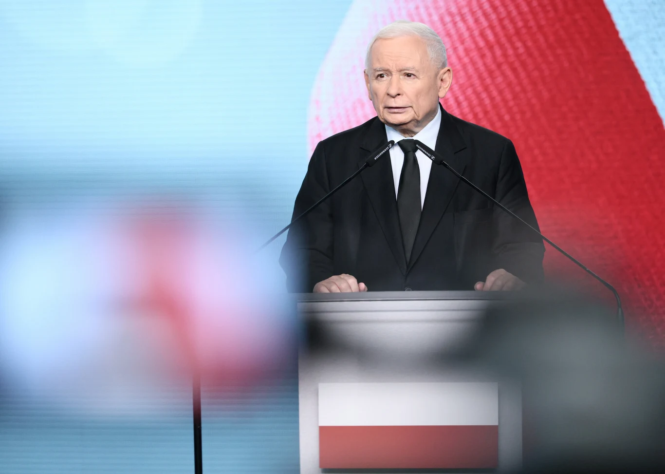 Starszy mężczyzna w garniturze wygłaszający przemówienie przy mównicy z flagą Polski, na tle dużego ekranu z motywem biało-czerwonym.