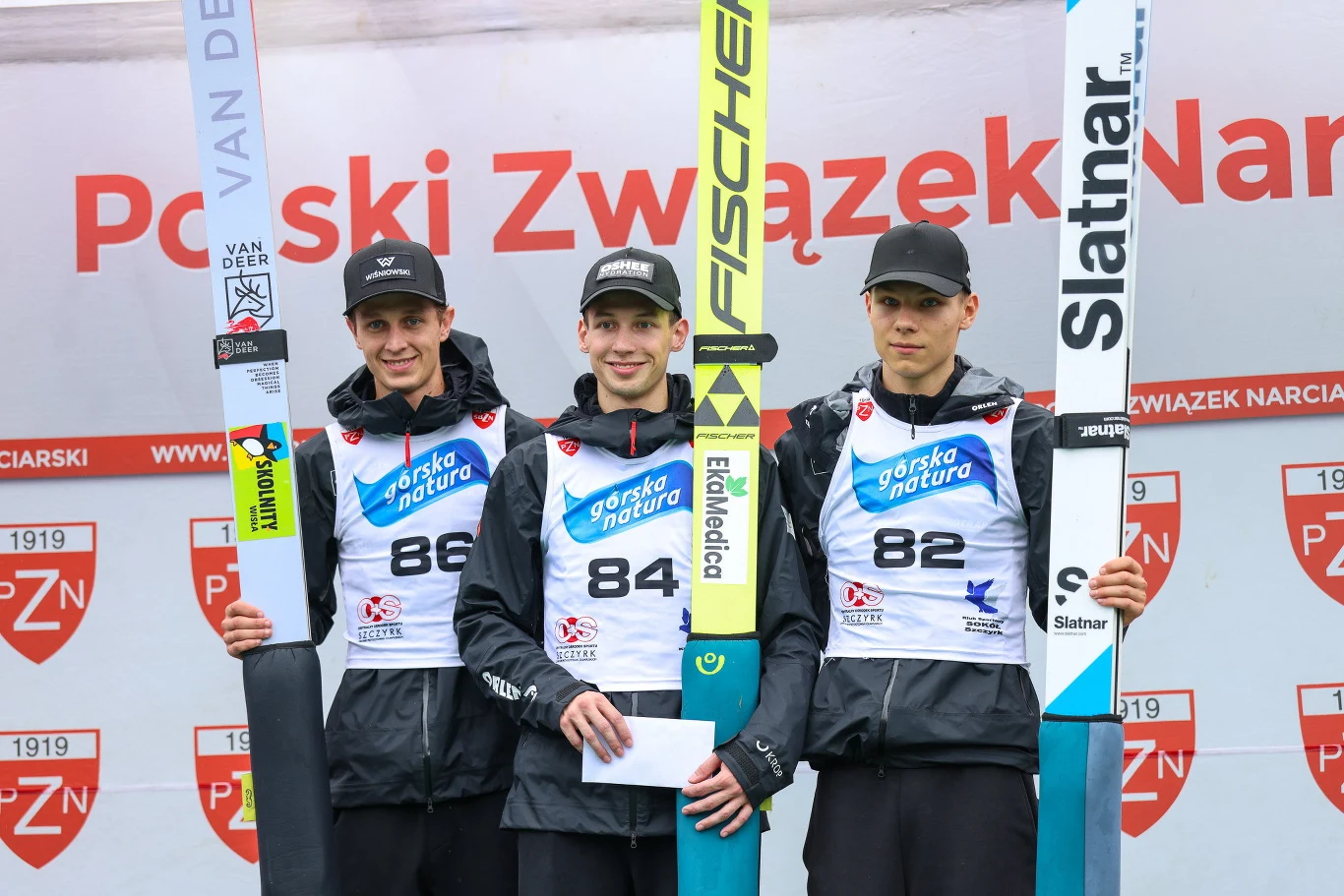 Trzech młodych skoczków narciarskich w sportowych strojach, stojących na podium z nartami w dłoniach i numerami startowymi na piersiach, na tle baneru organizatora zawodów.