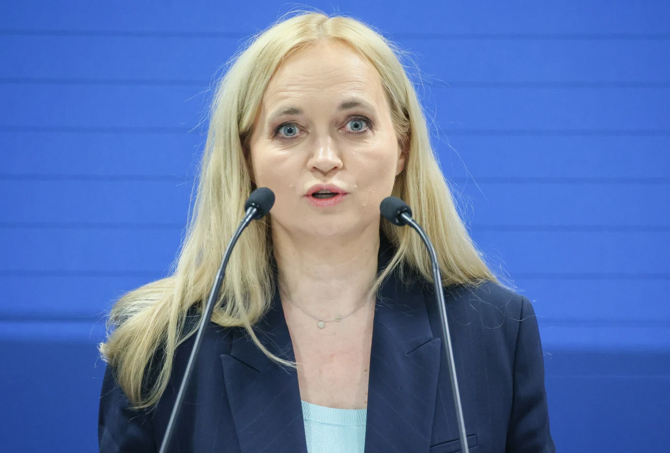 Minister zdrowia Jolanta Sobierańska-Grenda Minister zdrowia Jolanta Sobierańska-Grenda w granatowej marynarce mówi do mikrofonów na niebieskim tle.