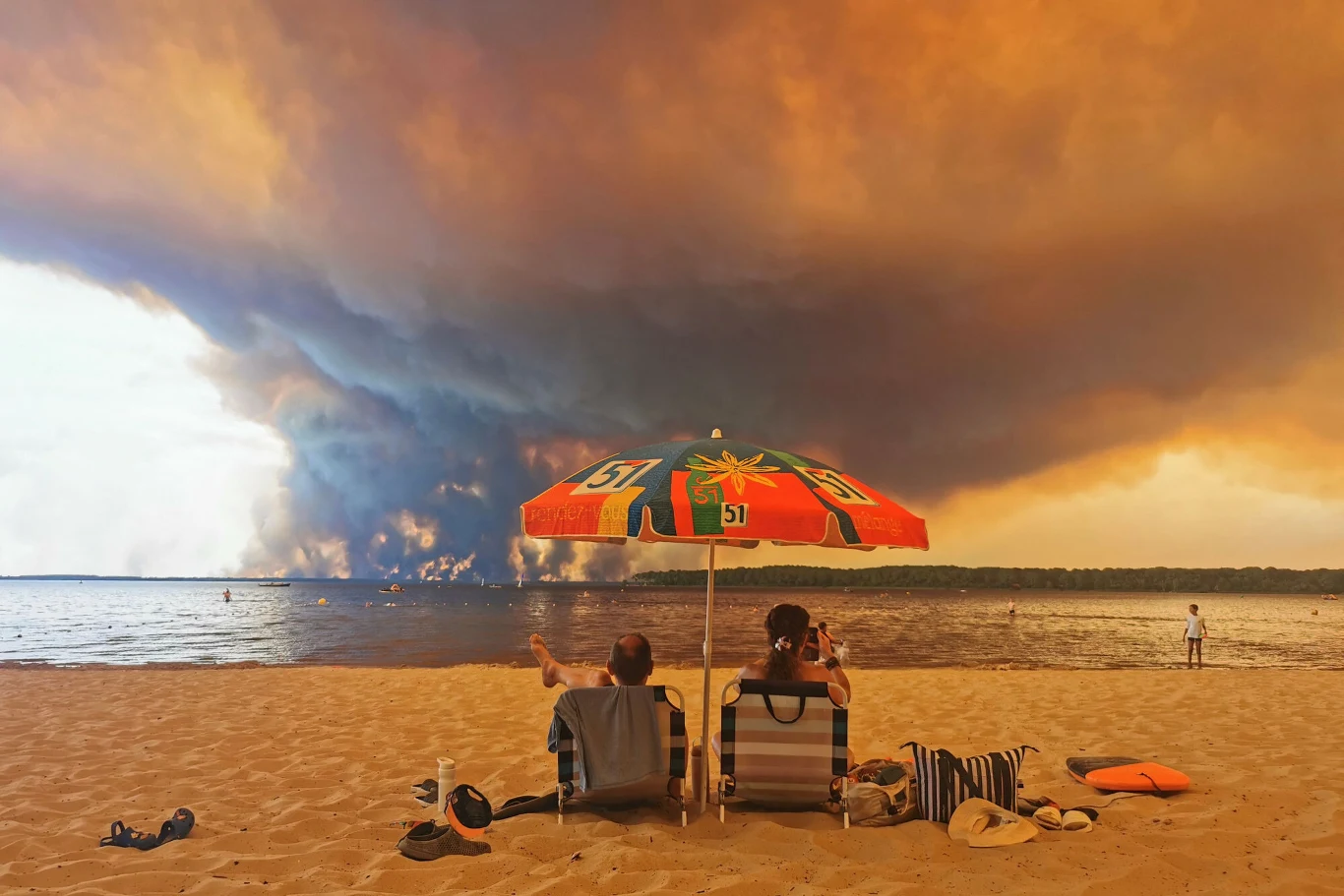 Dwie osoby siedzące na plaży pod kolorowym parasolem, na horyzoncie rozległa chmura dymu unosząca się nad lasem po drugiej stronie jeziora.