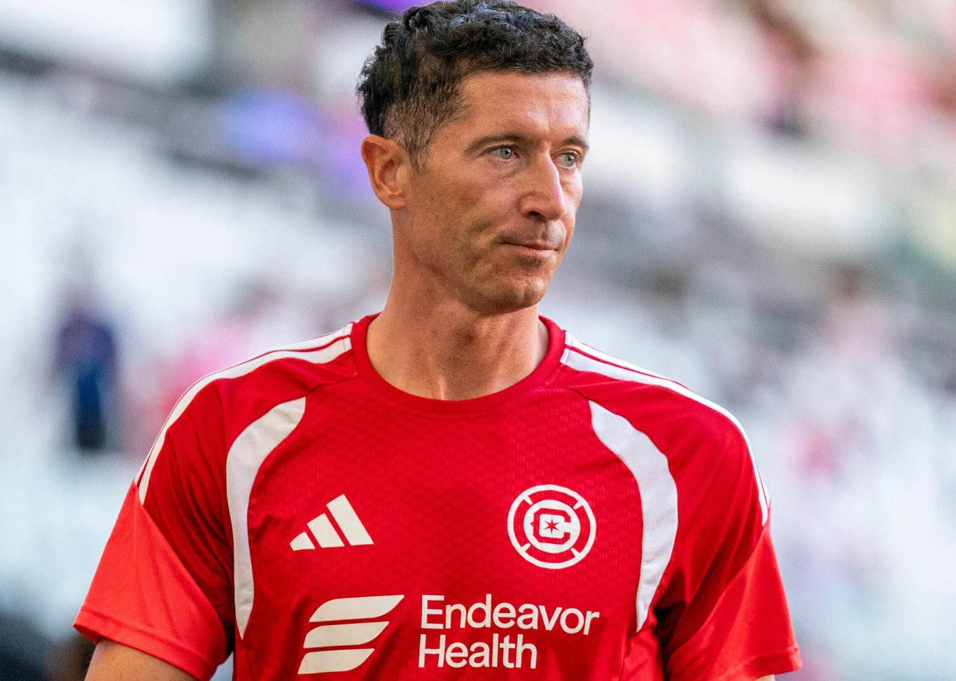Robert Lewandowski w barwach Chicago Fire Mężczyzna w sportowej, czerwonej koszulce z białymi nadrukami i logo klubu piłkarskiego stoi na stadionie, patrząc przed siebie.