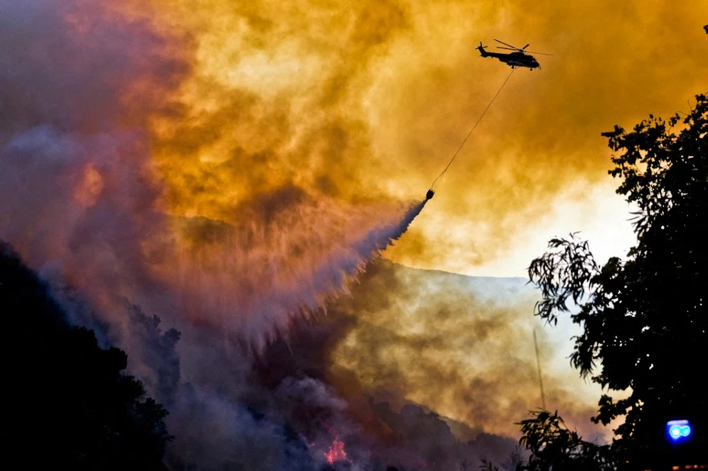Francja w ogniu. Fala pożarów paraliżuje region Bordeaux. Trwa akcja gaśnicza Helikopter gaśniczy zrzuca wodę na płonący las, wokół gęsty dym i intensywne płomienie, na pierwszym planie zarys drzew.