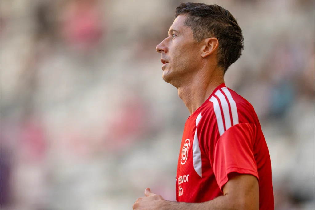 Robert Lewandowski Piłkarz w czerwonej koszulce treningowej z białymi paskami na ramionach, widoczny w półprofilu na tle rozmytego stadionu.