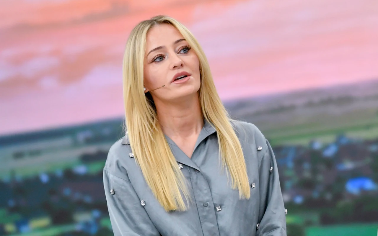 Kobieta o długich blond włosach, ubrana w jasnoniebieską koszulę z ozdobnymi guzikami, stoi na tle panoramicznego krajobrazu z polami i zabudowaniami, z delikatnie różowym niebem w tle.