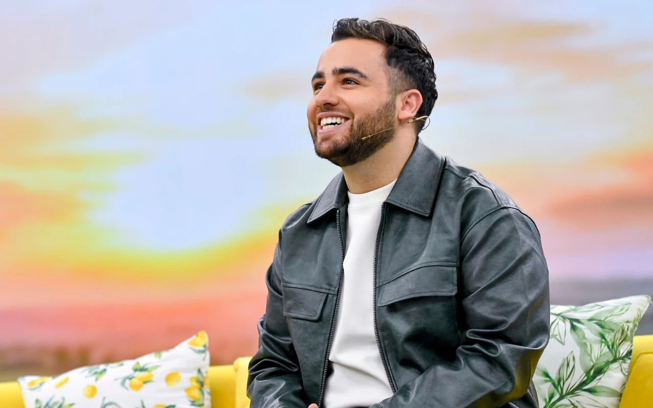 Sargis w "halo tu polsat" Uśmiechający się mężczyzna w ciemnej, skórzanej kurtce siedzi na żółtej sofie ozdobionej poduszkami z motywem cytryn i zielonych liści, w tle kolorowy zachód słońca.