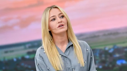 Kobieta o długich blond włosach ubrana w szarą koszulę z ozdobnymi guzikami, z mikrofonem nagłownym, znajduje się na tle pastelowego nieba i rozległego krajobrazu wiejskiego.