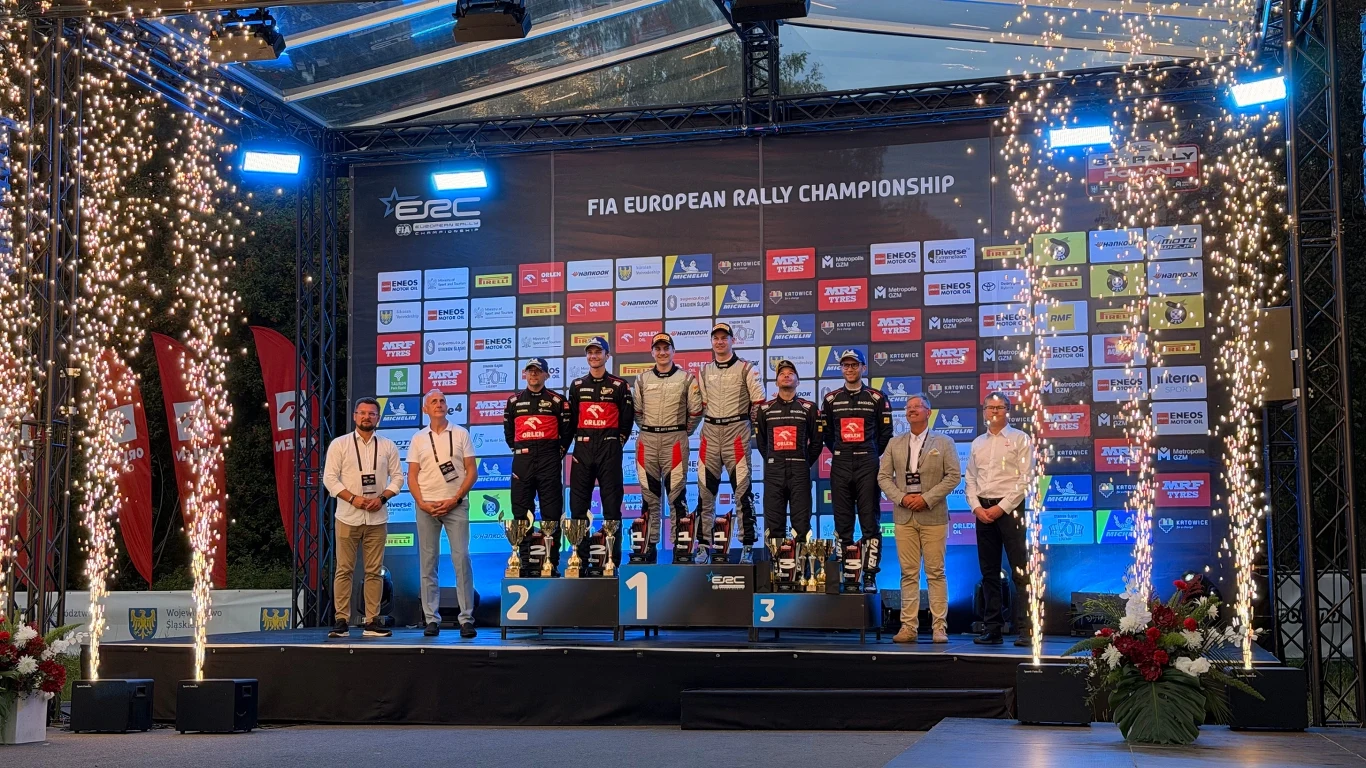 Zwycięzcy i laureaci rajdu stoją na podium FIA European Rally Championship, otoczeni dekoracjami świetlnymi i skąpani w blasku fajerwerków, z tyłu widoczna jest ścianka sponsorów i organizatorów.
