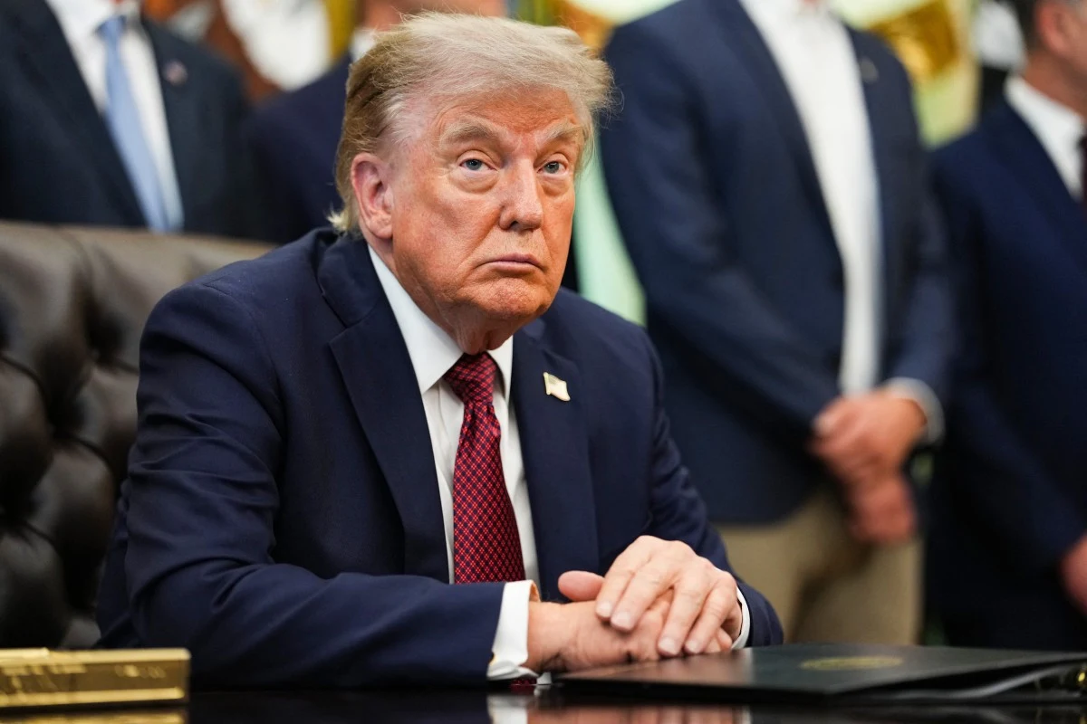 Prezydent USA Donald Trump Donald Trump w granatowym garniturze i czerwonym krawacie siedzi przy biurku, w tle osoby w formalnych strojach.