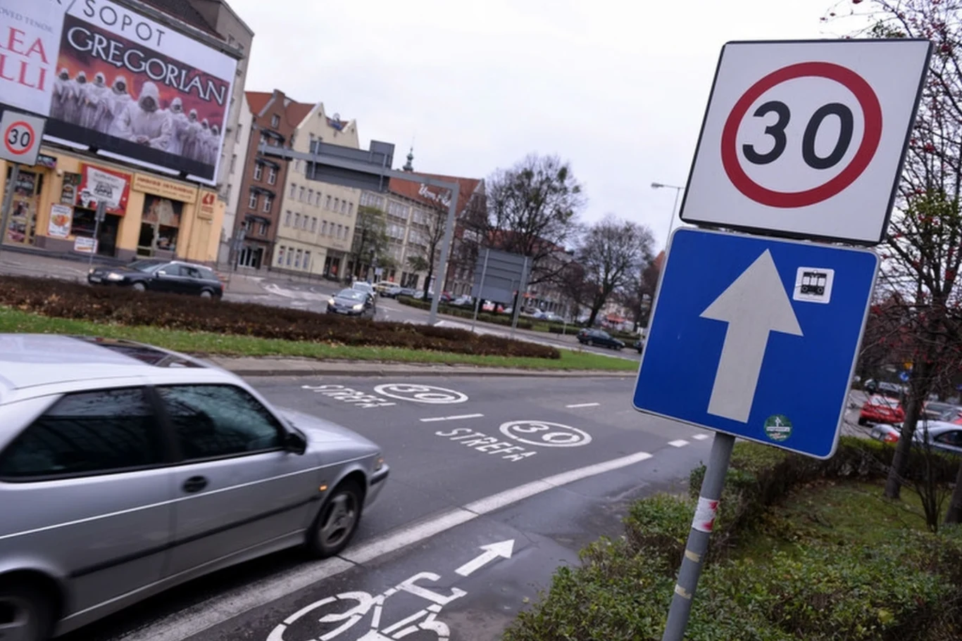 Srebrny samochód osobowy poruszający się po ulicy, znaki drogowe ograniczające prędkość do 30 km/h oraz oznaczenie drogi rowerowej, w tle kamienice i billboard reklamowy.