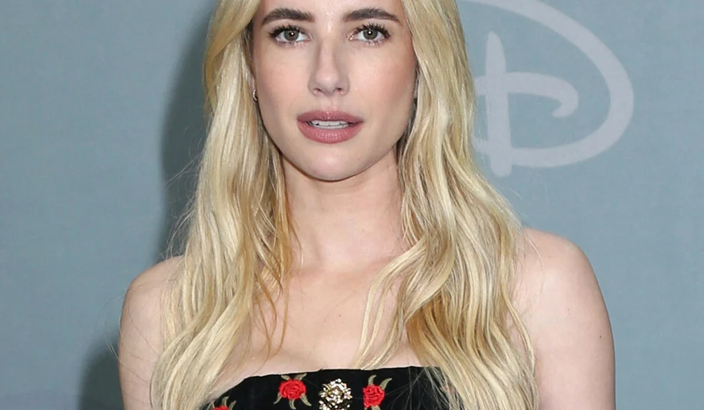 Emma Roberts Emma Roberts w czarnej sukience w haftowane kwiaty na tle niebieskiej ściany z logo.