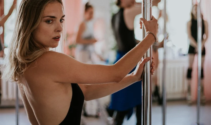 Kobieta o jasnych włosach trzyma drążek do pole dance, skupiona na ćwiczeniu w sali treningowej, w tle rozmazane sylwetki innych osób przy drążkach.
