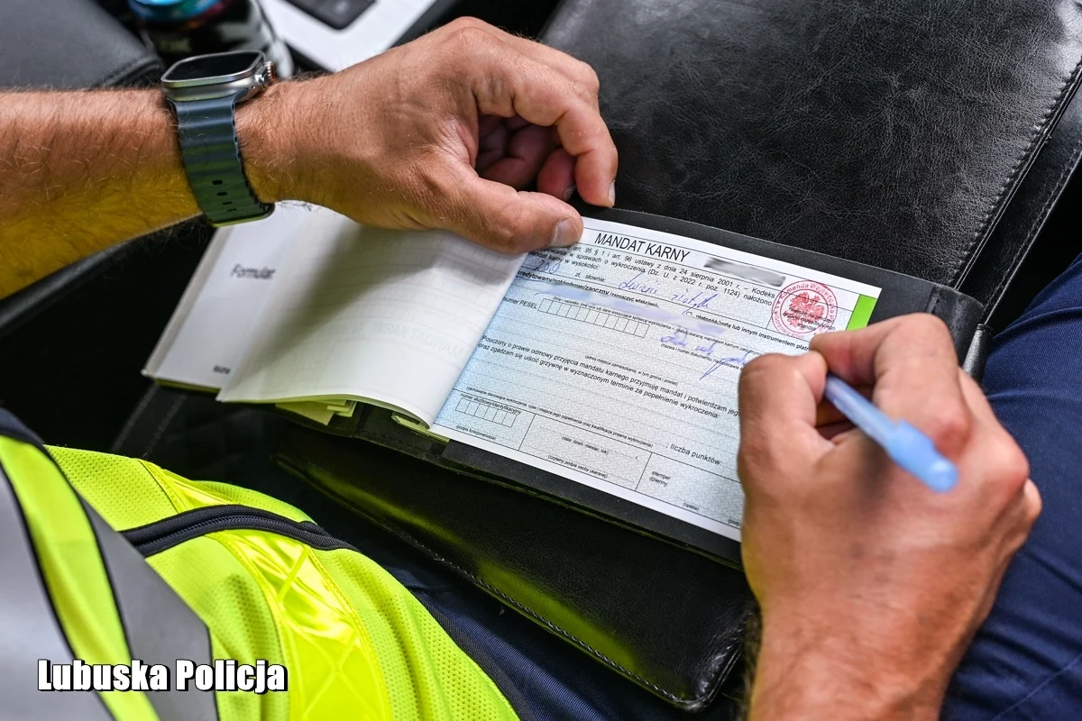 Policjant w żółtej kamizelce wypisuje mandat karny na specjalnym blankiecie, trzymając długopis nad czarną teczką.