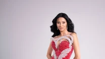 Kobieta z długimi, ciemnymi włosami w eleganckiej, dopasowanej czerwonej sukni wieczorowej z błyszczącymi zdobieniami i wycięciem przy dekolcie, stojąca przy białych stopniach na neutralnym tle.
