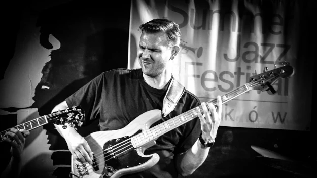 Mężczyzna gra na gitarze basowej podczas koncertu jazzowego, widoczne intensywne zaangażowanie w grę.