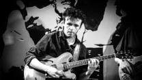Mężczyzna grający na gitarze elektrycznej, skoncentrowany na muzyce, z towarzyszącym basistą w tle.