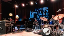 Trzech muzyków na scenie podczas koncertu jazzowego: pianista przy fortepianie, perkusista grający na zestawie perkusyjnym oraz instrumentalista siedzący pośrodku, w tle widoczne logo Summer Jazz Festival Kraków na ciemnej kurtynie.