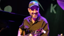 Mężczyzna z brodą i tatuażami grający na gitarze elektrycznej, uśmiechnięty, ubrany w ciemną koszulę i czapkę z daszkiem, oświetlony światłami sceny.