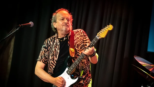 Starszy mężczyzna grający na gitarze elektrycznej na scenie, ubrany w koszulę w zwierzęcy wzór, na tle ciemnych zasłon i oświetlenie koncertowe.