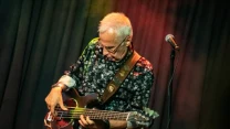 Starszy mężczyzna z siwymi włosami gra na gitarze basowej, ubrany w ciemną koszulę w kwiatowy wzór, obok mikrofon na statywie na tle ciemnej, scenicznej zasłony.