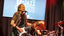 Mężczyzna grający na gitarze elektrycznej i śpiewający do mikrofonu na scenie podczas koncertu, w tle perkusista i baner festiwalu jazzowego.