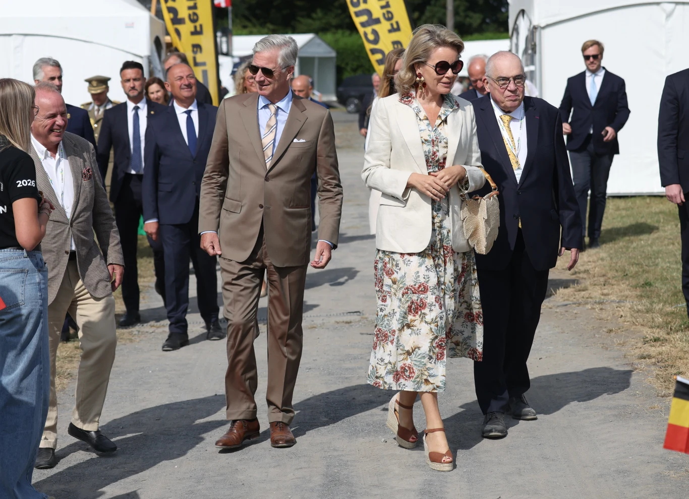 Elegancko ubrana kobieta w kwiecistej sukience i jasnej marynarce oraz mężczyzna w beżowym garniturze kroczą razem alejką, otoczeni przez grupę towarzyszących im osób w formalnych strojach.