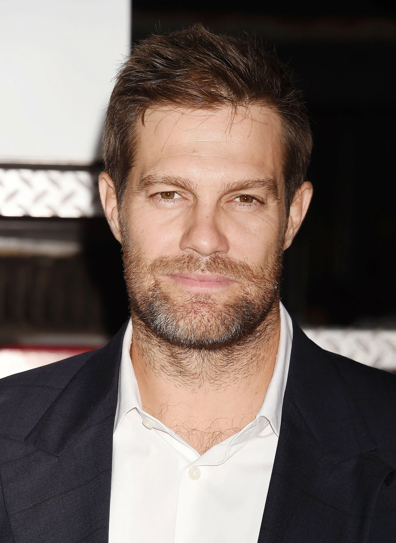 Geoff Stults Mężczyzna z krótkimi brązowymi włosami i zarostem, ubrany w białą koszulę i ciemną marynarkę, patrzy wprost w obiektyw.