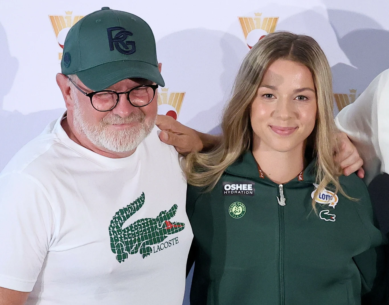 Mężczyzna w okularach i zielonej czapce z daszkiem obejmuje młodą kobietę ubraną w sportową bluzę z logo Lacoste i naszywkami sponsorów, oboje uśmiechają się do aparatu.