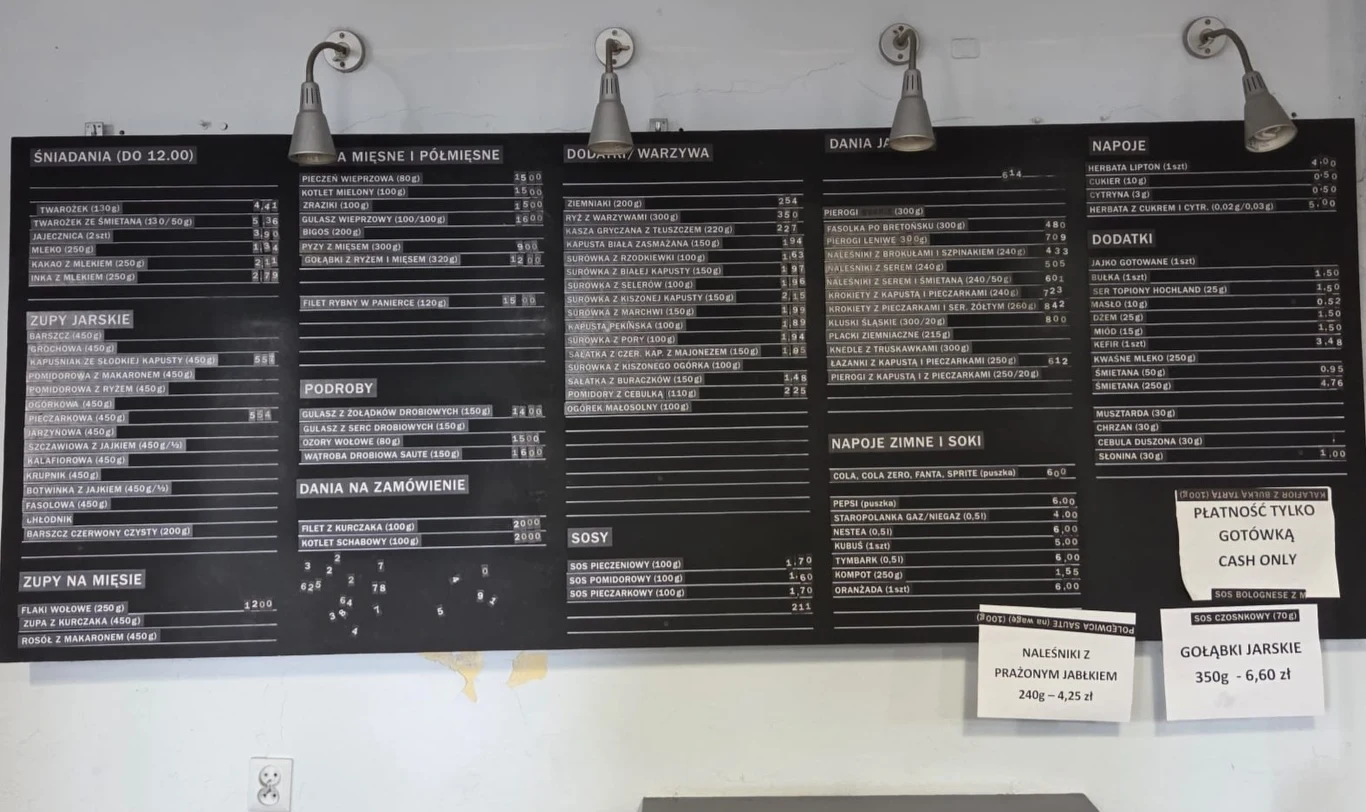 Duża czarna tablica z białym tekstem pełni funkcję menu, podzielonego na kategorie takie jak śniadania, zupy, mięsa, dania warzywne, napoje i dodatki; po prawej stronie widoczne są dodatkowe kartki informujące o formie płatności oraz aktualnej cenie za...