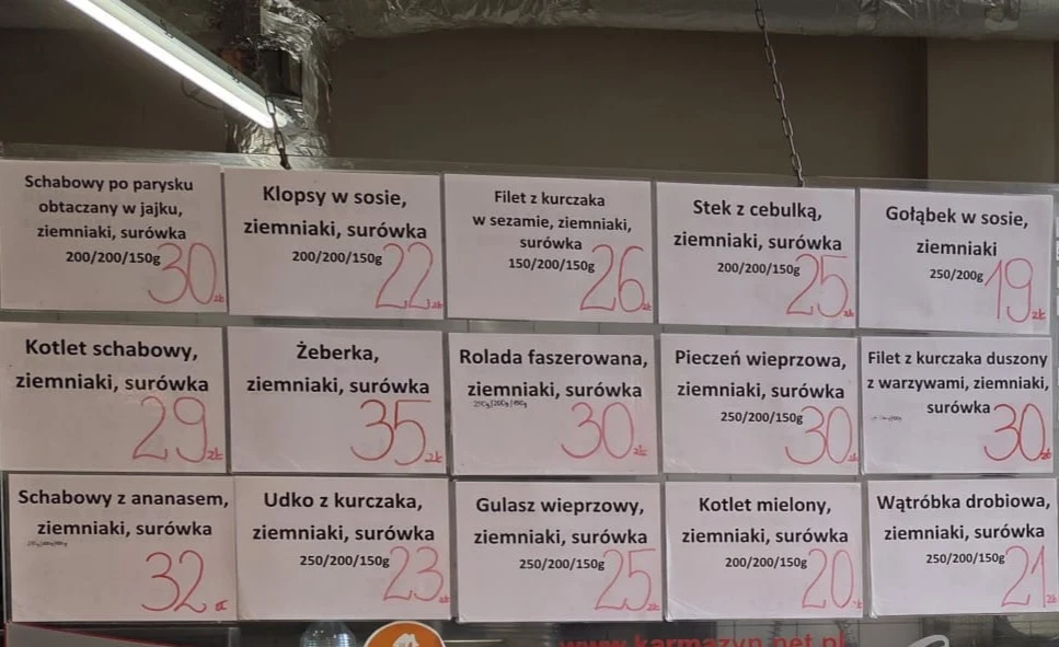 Tablica z ofertą dań obiadowych wypisanych na białych kartkach, obejmujących różne rodzaje mięs z dodatkiem ziemniaków i surówki, z wyraźnie zaznaczonymi cenami przy każdym zestawie.