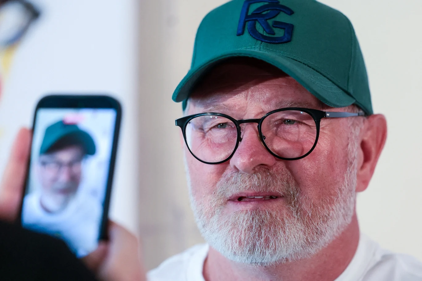 Jaroslav Machovsky Starszy mężczyzna z siwą brodą i okularami, w zielonej czapce z daszkiem, patrzy przed siebie, podczas gdy ktoś robi mu zdjęcie telefonem komórkowym, na ekranie którego widoczny jest jego portret.