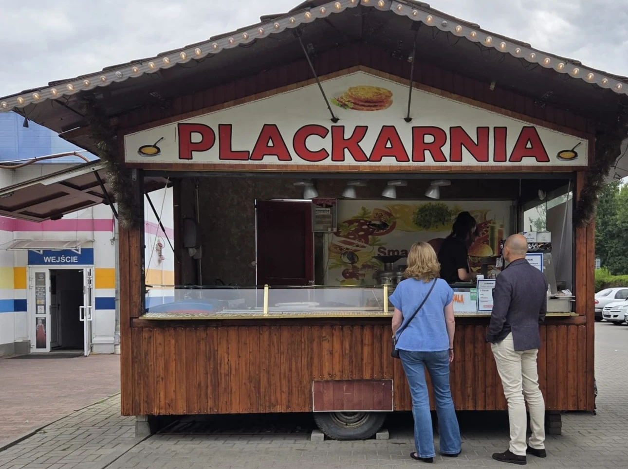 Drewniana budka gastronomiczna z dużym szyldem „Plackarnia”, przed którą stoją dwie osoby oczekujące na zamówienie.