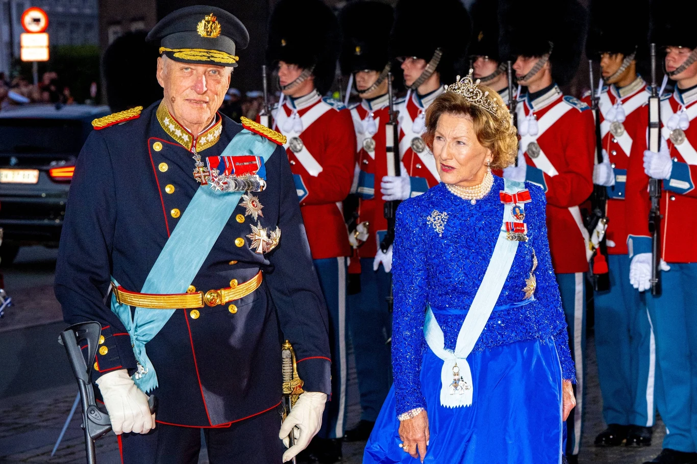 Król Harald V i królowa Sonja Starszy mężczyzna w galowym mundurze wojskowym z medalami i starsza kobieta w niebieskiej wieczorowej sukni oraz diademie, oboje noszą szarfy odznaczeniowe, idą razem przed szeregiem żołnierzy w historycznych mundurach z wysokimi czarnymi czapkami i cz...