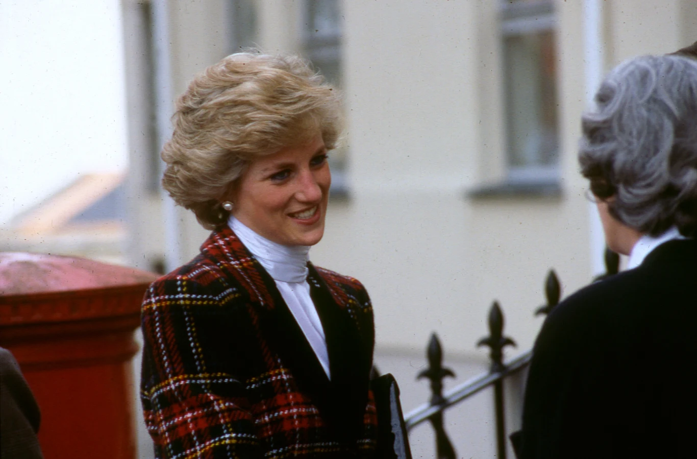Księżna Diana w 1980 r. Kobieta z krótkimi blond włosami ubrana w elegancką marynarkę w kratę i białą bluzkę rozmawia z drugą osobą na zewnątrz przy metalowym ogrodzeniu.
