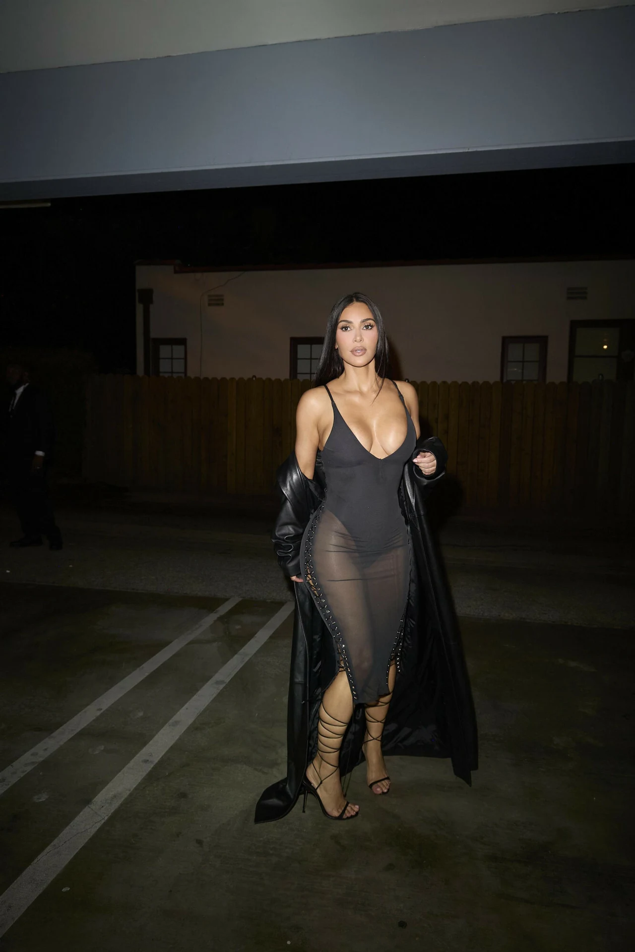 Kim Kardashian wie, jak podkreślić swoją figurę i wyglądać niezwykle kobieco Kobieta w długim, czarnym płaszczu i przezroczystej sukni z głębokim dekoltem, stojąca na parkingu wieczorową porą.