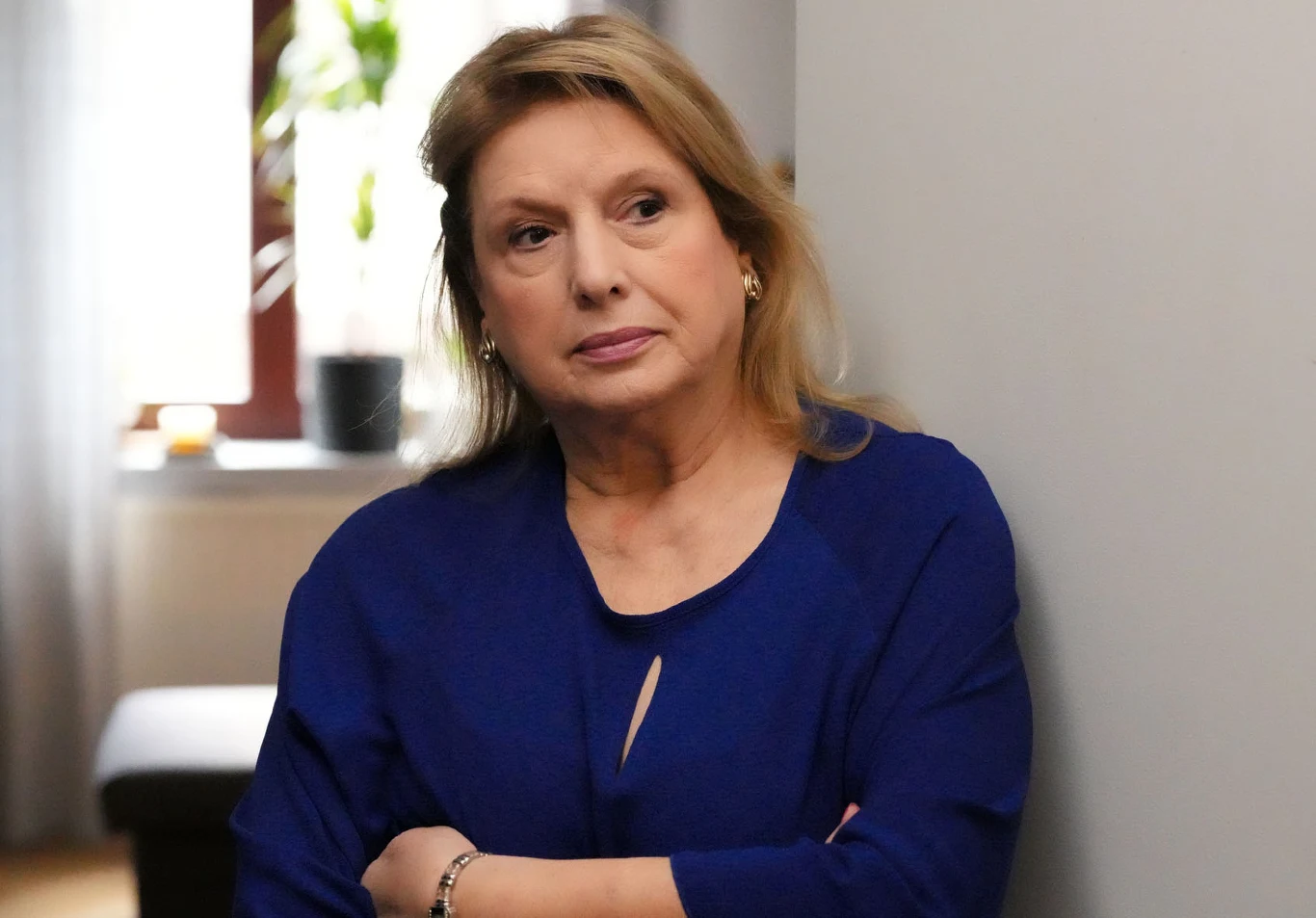 Kobieta w średnim wieku z blond włosami ubrana w niebieską bluzkę stoi oparta o jasną ścianę, z założonymi rękami i zamyślonym wyrazem twarzy.