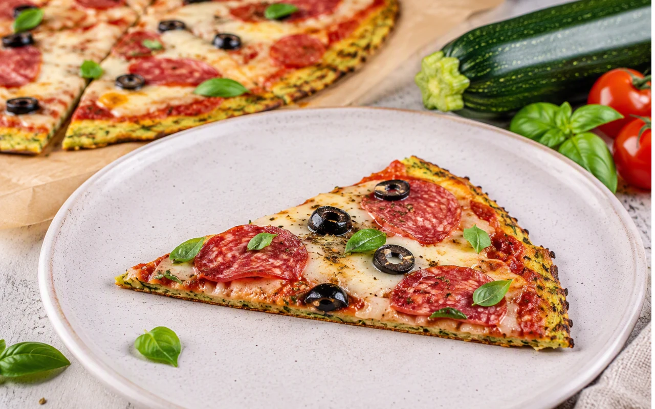 Trójkątny kawałek pizzy z plasterkami salami, czarnymi oliwkami, roztopionym serem i listkami świeżej bazylii na jasnym talerzu, obok warzywa: cukinia i pomidory koktajlowe.