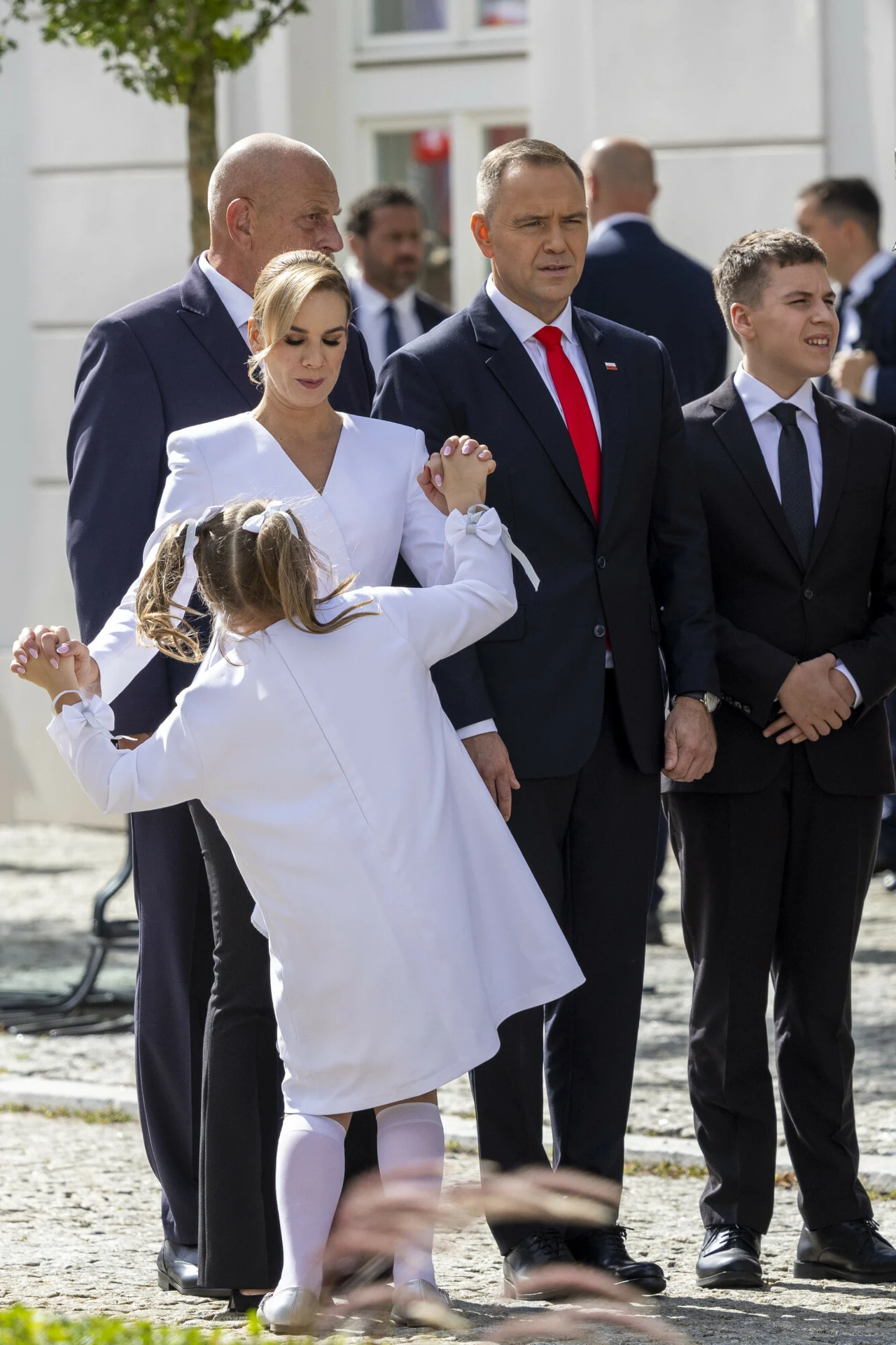 Grupa elegancko ubranych osób stoi razem na zewnątrz, w centrum uwagi znajduje się mężczyzna w garniturze z czerwonym krawatem oraz kobieta w białej sukni, przed nimi mała dziewczynka w białym stroju energicznie się obraca.