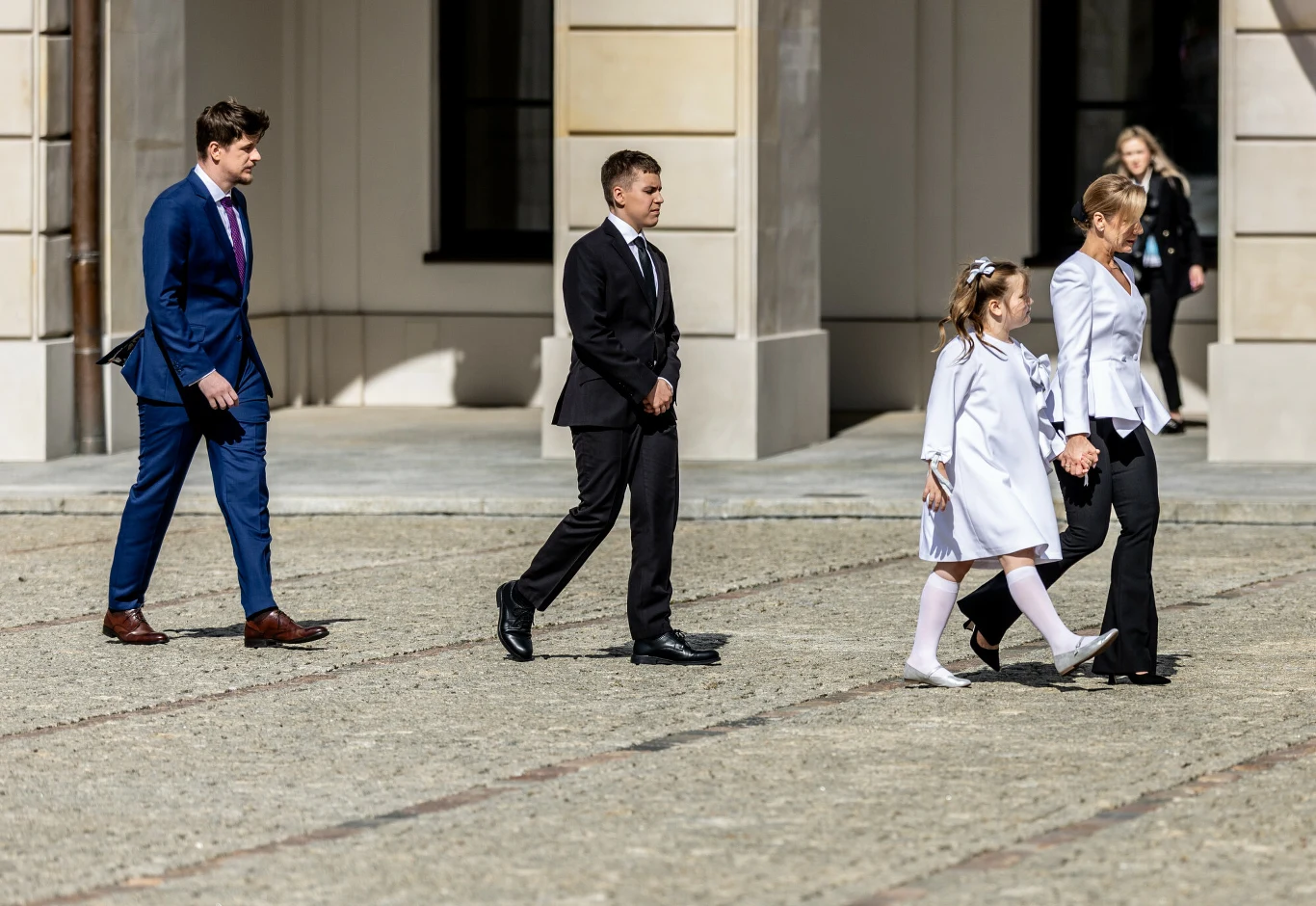 Grupa osób ubranych elegancko, dwie dziewczynki ubrane na biało oraz troje dorosłych, przechadza się po jasnym bruku przed dużym budynkiem.