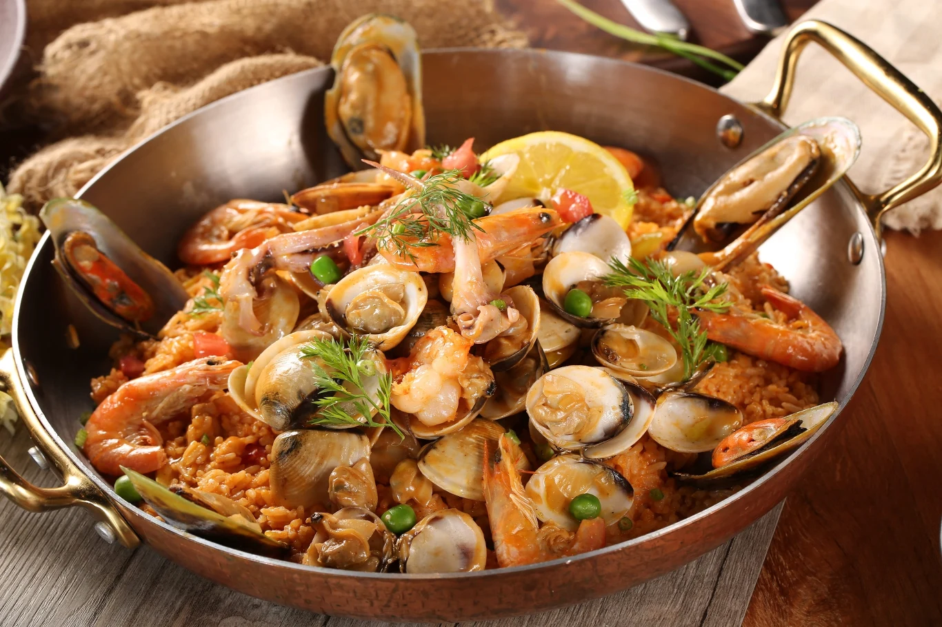 Hiszpańska paella z owocami morza zachwyca wyglądem i smakiem. Paella z owocami morza, krewetkami, małżami i cytryną w metalowej patelni, udekorowana ziołami.
