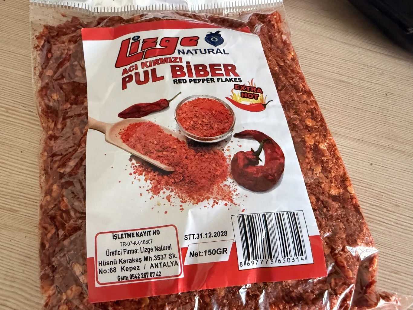 Opakowanie przyprawy w postaci płatków ostrej czerwonej papryki z widoczną etykietą zawierającą nazwę produktu, zdjęcia papryki i potrawy, a także szczegóły dotyczące wagi i producenta.