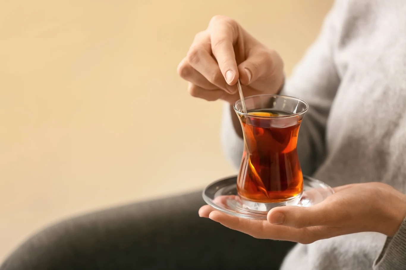 Szklanka z herbatą umieszczona na szklanym spodku, trzymana w jednej ręce, a drugą ręką osoba miesza napój łyżeczką.
