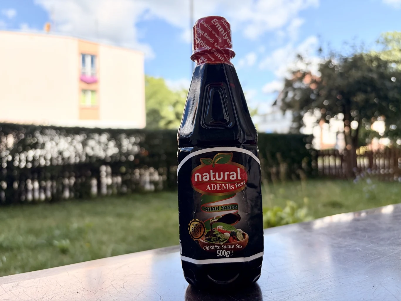 Butelka sosu sojowego natural marki Adem's tet z czerwonym korkiem umieszczona na stole, w tle rozmyty widok ogrodu i budynku mieszkalnego.