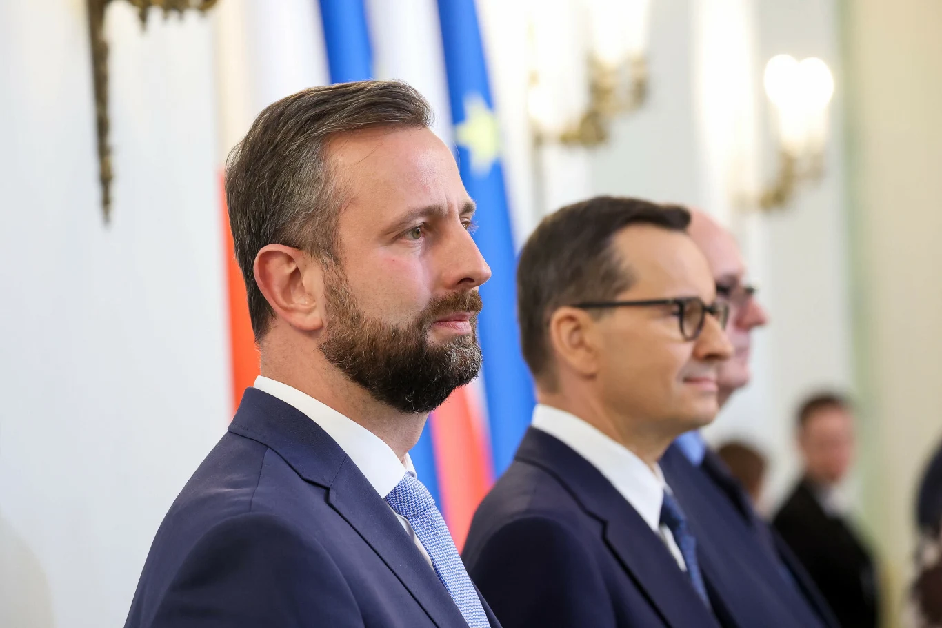 Trzech mężczyzn w garniturach stoi w szeregu na tle flag Polski i Unii Europejskiej w eleganckim, jasnym wnętrzu.
