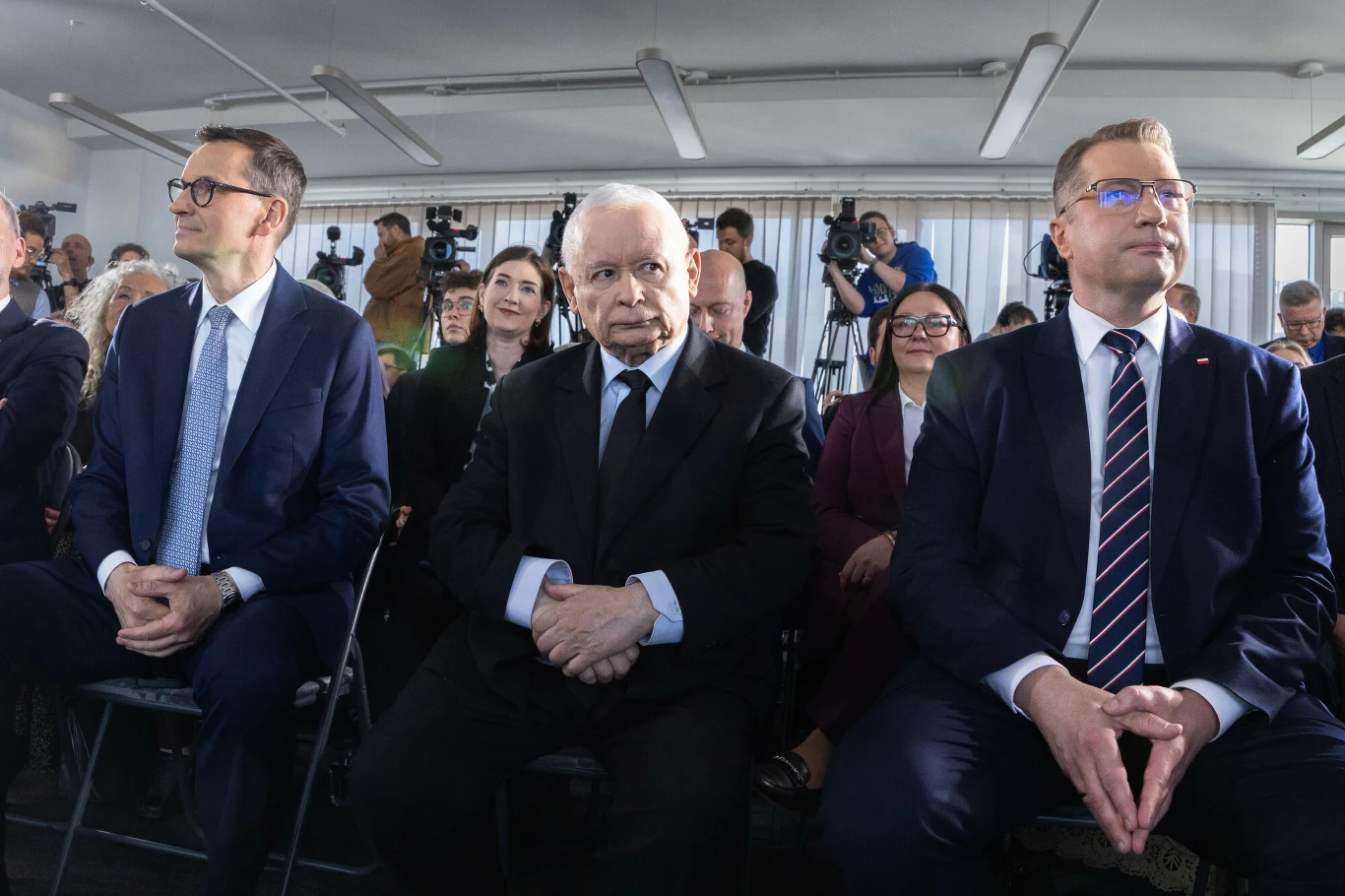Trzech elegancko ubranych mężczyzn siedzi w pierwszym rzędzie podczas oficjalnego wydarzenia, a za nimi widoczni są uczestnicy i fotoreporterzy z aparatami.