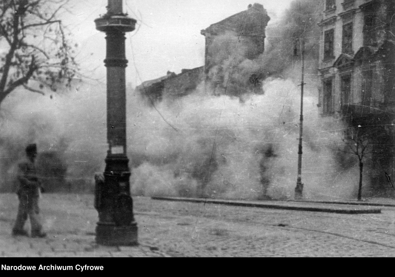 Gęsta chmura dymu unosząca się nad ulicą w centrum miasta, zniszczone budynki w tle, samotna postać przechodząca obok latarni na pierwszym planie.